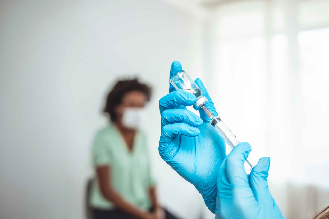 Imagem ilustrativa da matéria: Enel unidos pela vacina. Possui mãos com luvas azuis segurando uma seringa e uma dose da vacina, com uma mulher sentada ao fundo